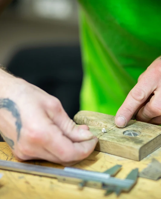A close-up of a tattooed craftsman’s hands assembling a jewellery piece on a wooden workbench, using a small gemstone and delicate wire. A close-up of a tattooed craftsman’s hands assembling a jewellery piece on a wooden workbench, using a small gemstone and delicate wire.